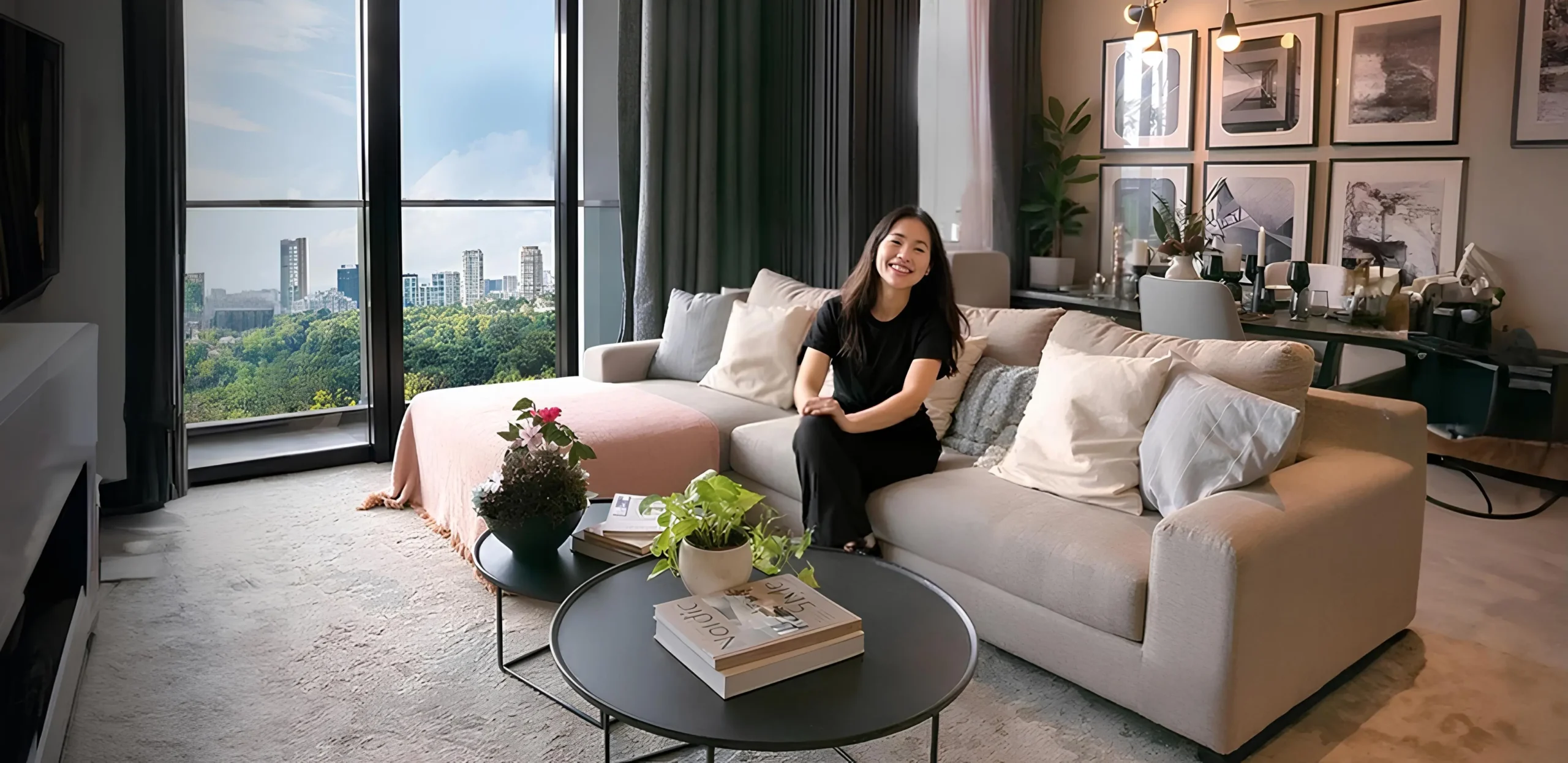 Seorang wanita tersenyum duduk di ruang tamu show unit EleVee Residences dengan pemandangan kota dan hutan kota dari jendela besar.
